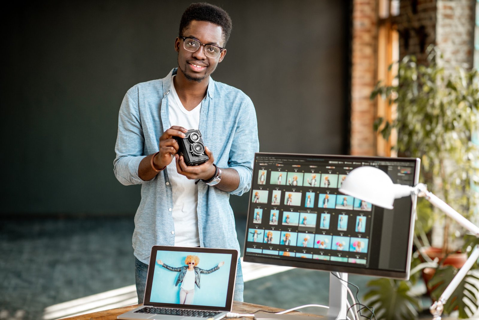 portrait-of-a-young-african-ethnicity-photographer-standing-with-retro-camera-near-the-working-place-with-computers-in-the-studio