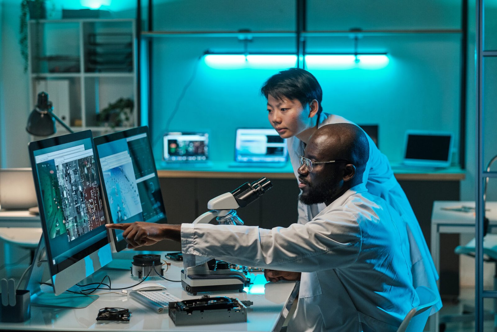 two-young-intercultural-professionals-in-whitecoats-discussing-microchip-scheme-while-one-of-them-pointing-at-computer-screen